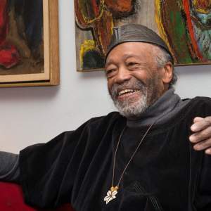 Otto Neals smiles on a couch surrounded by artwork. He is Black with a greying beard, and wears a leather cap and a necklace with a cowrie shell pendant.