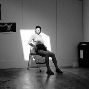 Black and white photo of the artist Daniela Leal, sitting on a step stool in a square of light, with photographs on white wall behind her