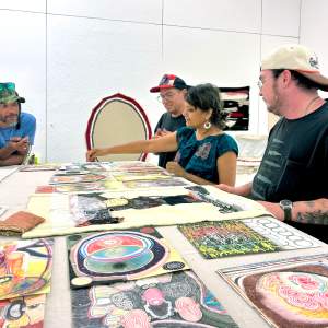 4 people of various age and appearance gather around a work table covered with colorful abstract artworks on paper and fabric..