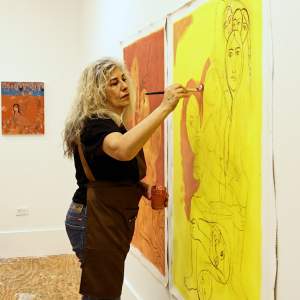 Samira Abbasy stands in a studio painting abstracted figures onto a large sheet paper tacked to the wall. She is a woman of Iranian decent with long blond hair and medium light skin tone, wearing an apron, jeans and a tshirt.