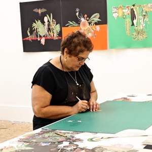 Anila Quayyum Agha cuts paper at work table in her art studio. She has short dark hair and glasses, wears a black shirt and necklace, and has a medium light skin tone.