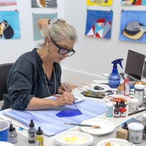 Sarah paints a deep blue geometric shape onto light blue-washed paper, on a table surrounded by small containers of paints, dyes, and brushes. Behind her are a group of geometric abstract works on paper clipped to the wall. She has fair skin, light blonde-grey hair, and dark framed glasses.