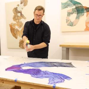 A man with light skin, blonde short hair, and glasses sprays a wavy, delicate piece of silk stretched out on a table in front of him.