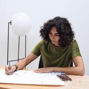 Diane Appaix-Castro sits at a table drawing on a large piece of paper in a white studio. She has medium-light skin and dark curly hair, and wears a green shirt.