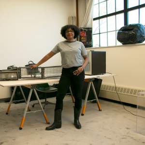 Ash Arder, a Black woman with medium-dark skin tone, dark curly chin length hair, stands in an art studio next to a table. A detached white Cadillac car door leans on a wall.