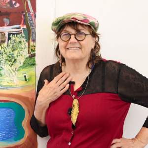 Olive Ayhens, a white woman with chestnut hair under a floral newsboy hat, smiles with her left hand on her chest. She wears round glasses, a red and black shirt, and a necklace with a large gold feather.