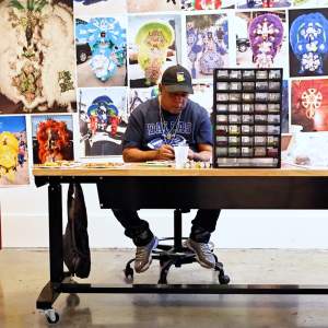 Big Chief Pie, a Black man with medium skintone wearing a baseball hat and Dallas cowboys shirt, sews beadwork at a worktable in front of photographs of Black Masking Indian regalia.