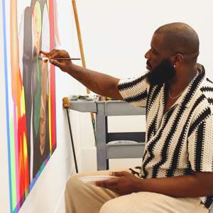 Anthony Badon sits and paints onto a canvas on the wall. He is a Black man with dark skin tone, a black beard and wears a striped knit shirt.