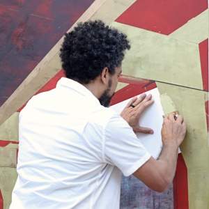 Rogelio Baez Vega scrapes gold paint onto a large graphical canvas. His back to us, he has dark curly greying hair, and medium skin tone.