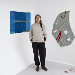 Teresa Baker, a Native American and German American woman with medium-brown skin and long black and gray hair in a braid, stands with her hands in her pockets between two abstract wall works, hung in the corner of a white room.