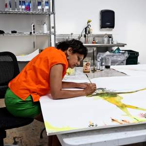 Rina Banerjee, an Indian-American woman with medium skintone and short black hair, leans over a large table working on a figurative watercolor.