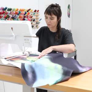 Sophia Belkin, a white person with long dark hair and light skin, works at an embroidery machine with a multicolor piece of fabric.