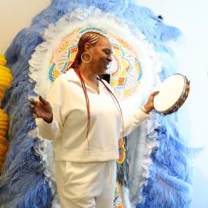 Big Queen Kelly smiles and shakes a tambourine in front of a large feathered regalia suit. She is a Black woman with cornrows of red braids, and wears hoop earrings and a matching white tracksuit.