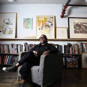 Michaela Pilar Brown sits in a grey tweed chair in a room lined with low bookshelves and framed artwork on a shelf above. She is a black woman with a shaved head and wears chunky black glasses, and wears large rings, a black jumpsuit, and converse sneakers.