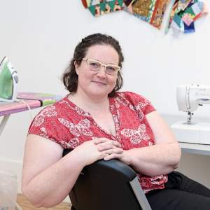Ellen Bull, a white woman with chin length brown hair, sits resting her arm on the back of a chair in front of a table with a sewing machine in an art studio.