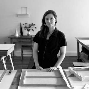 In a black and white photo, Victoria Burge, a white woman with light skin tone and long dark hair, stands at a work table in her studio, stretching a canvas that is upside-down on the table in front of her. She smiles slightly at the camera and wears a dark zippered jumpsuit and half-moon earrings.