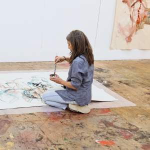 Virginia Candler, a white woman with medium-light skin tone and long salt-and-pepper hair, kneels on the floor working on a large abstract watercolor.