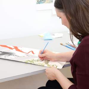 At a gray table, Carlson paints details of a cypress tree on paper with a fine brush. She has long brown hair, light-toned skin, and wears a burgundy sweater.