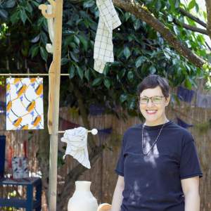 Dawn Cerny, a white woman with short brown hair and tortoiseshell glasses, smiles for the camera in her backyard next to a large wooden sculpture with pegs that have dish cloths and other fabric hanging from them and a piece of pottery nestled into a tabletop.
