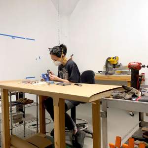 Peggy Chiang sits at a worktable in her studio, wearing a respirator and filing something that is clamped to the table. She has black hair and medium skin tone and is surrounded by power tools and other sculpture-making materials.