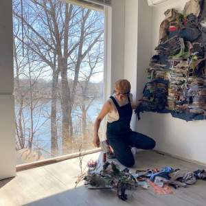 Ruby Chishti kneels in the corner of her studio, a room with white walls and wooden floors, with a window behind her showcasing a view of a lake and trees. Ruby is a Pakistani-American woman with medium light skin and short dark hair. She wears overalls, white tank top, and brown hat and works with fabric strips on a dress-like sculpture that she is assembling on the wall.