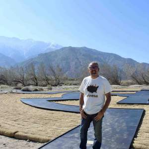 Gerald Clarke stands on a land art installation of a black pathway stretching behind him, installed in Palm Springs desert with  mountains visible in the background under a clear blue sky is a citizen of the Cahuilla Band of Indians, and has a medium skin tone and a salt-and-pepper goatee, and his hair is pulled back, wand wears a tshirt with outline of the US reading “amnesia”.