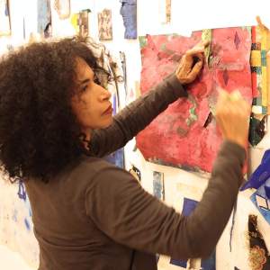 Yanira glances to the right as she works on a textured magenta paper pinned to the wall, which has a number of scraps of patterned and textured paper. She has medium-light skin, and dark brown curly hair.
