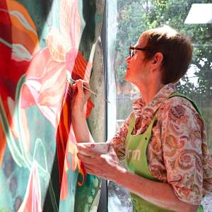 Elisabeth Condon, a white woman with short red hair wearing glasses and a floral shirt, paints on a large floral painting as light streams in.