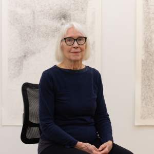 Cecily Cottingham sits in a bright white studio in front of large drawings. She is a white woman with light skin tone, white shoulder-length hair and wears glasses and black clothing.