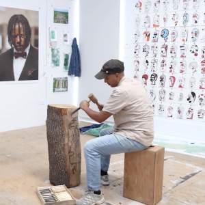 Jose Cotto sits on a wooden box, using a chisel and mallet to carve an abstract face from an upright log. Behind him are photographs and drawings pinned to a white studio wall.