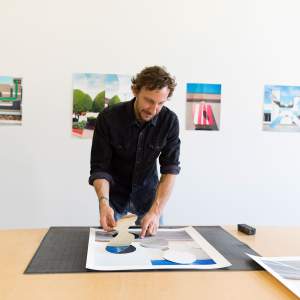 A white man with short curly brown hair and a beard leans over a work table, moving a piece of a cut photograph. Behind him are a row of photo collages.