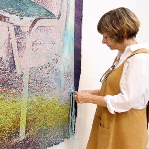 Artist Lauren Davis pulling blue threads from a distressed textile on a white wall