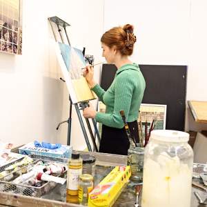 Paige DeVries paints on an easel in a white walled studio, in the foreground is a table with painting tools. She is a white woman with auburn hair, wearing a green shirt.