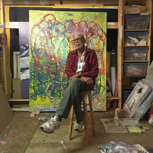 Bob Dilworth est assis sur un tabouret en bois dans un atelier aux étagères en pin et au sol en contreplaqué, une grande peinture figurative abstraite appuyée derrière lui. C'est un homme noir d'un certain âge, aux cheveux gris coupés court, qui porte une chemise à carreaux et des lunettes rondes en écaille.