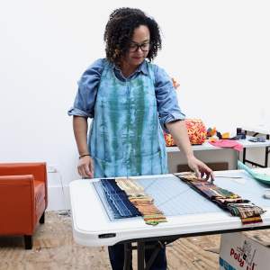 Addoley Dzegede works with swatches of cloth in a studio. She is a Black woman with medium light skin tone, dark shoulder length hair in tight curls, and wears a batik apron