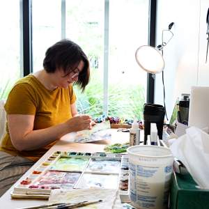 Dara Engler works at a studio table on an embroidery piece, surrounded by watercolor paints and other supplies. She is a white woman with light skintone and short straight brown hair, wearing a goldenrod shirt.