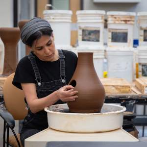 Raheleh Filsoofi, an Iranian-American woman with medium skin tone and dark hair, sits at a potter's wheel in a studio, with tables, plaster slabs, and buckets visible behind her. Wearing dark overalls and a black t-shirt, she cuts a hole into a bottle-shaped clay vessel on the wheel.