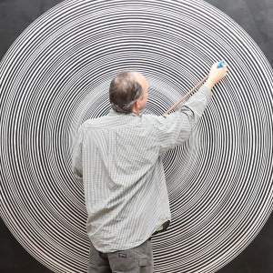 James Flynn, a white man with greying hair, works on a hanging artwork of concentric white circles on a black surface.
