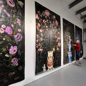 Four slightly glossy black panels hang side by side on a white wall, reaching nearly floor to ceiling. Sprays of similarly colored flowers arc out of vases at the bottom center of each. For instance, closest to us, insects and birds peek out from between purple-hued blossoms contained in a white and Delft-blue, round-bodied vase. The artist stands on a stepstool as she works on the third panel.