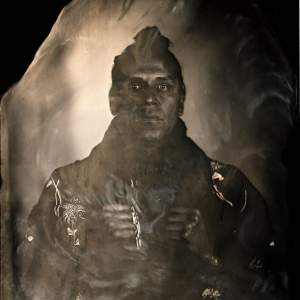 A double exposure platinum print photograph with Nicholas Galanin gazing directly. His face is darkened, dark hair in a bun, and he holds his lapels with his hands. A flower is carved into the surface on the left.