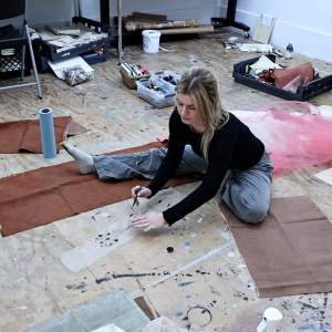 A white woman with blond hair sits on a paint splattered wooden studio floor, one leg straight and one bent painting with ink on gauze fabric.
