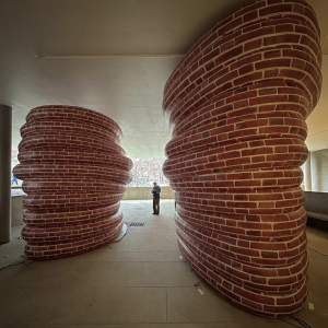Jacqueline Kiyomi Gork stands at a distance between two huge oblong pillars make from coiled tubes painted with a brick pattern.