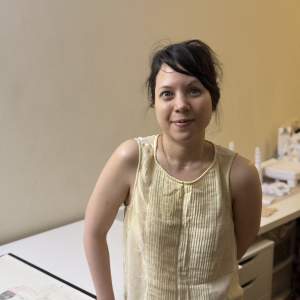 Emilie Louise Gossiaux leans against a drawing table in her home studio, smiling slightly at the camera. A petite woman of European and Indonesian descent, Gossiaux has dark brown hair pulled back in a messy bun, light tan skin, and wears a gauzy off-white tank top.