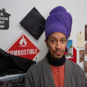 Against a studio wall with artwork and stickers tacked to the wall, André Leon Gray gazes at the camera. He is a Black man with medium dark skin tone and a mustache, and he wears a purple hair wrap, and gray and red layers.