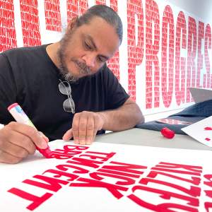 Joe Harjo sits at a work table in a gallery with a large mural artwork behind him, writing bold letters in red paint on a white board. Harjo is a Native American man with medium skin tone and long graying black hair pulled back, with a beard.