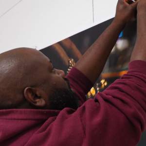 L. Kasimu Harris reaches his arms up to secure a photo on the wall in a burgundy hoodie, with a shaved head and beard