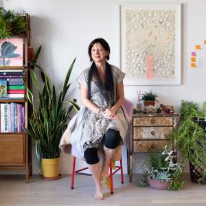 Ana Maria Hernando, an Argentinian woman with light skin tone and dark hair, sits on a red stool in the middle of a room full of plants, eclectic furniture, art and books. Smiling slightly at the camera, she wears a gray embroidered dress, a black tulle scarf, cropped black leggings, and no shoes.