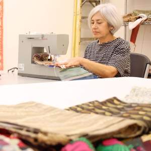 Elana works at a sewing machine. She has light skin and grey bobbed hair.