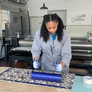 Hobbs leans over a woodcut with a repeating X pattern, holding with two gloved hands a large printing brayer, charged with royal blue ink. She is a Black woman with medium dark skin, with dark locs pulled back from her temples, and wears a gray jumpsuit.
