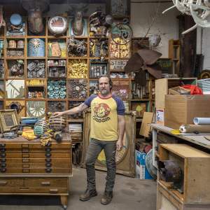 A white man with graying brown hair and a beard stands in an art studio loaded with materials, artwork, and boxes, resting his hand on a wooden sphere on top of a wooden flat file. He wears gray jeans, workbooks, and a yellow t-shirt reading “Creem.” Behind him is a grid of square shelves, each filled with a different assemblage, including a group of turquoise skulls, gears, paintings, and figurines.